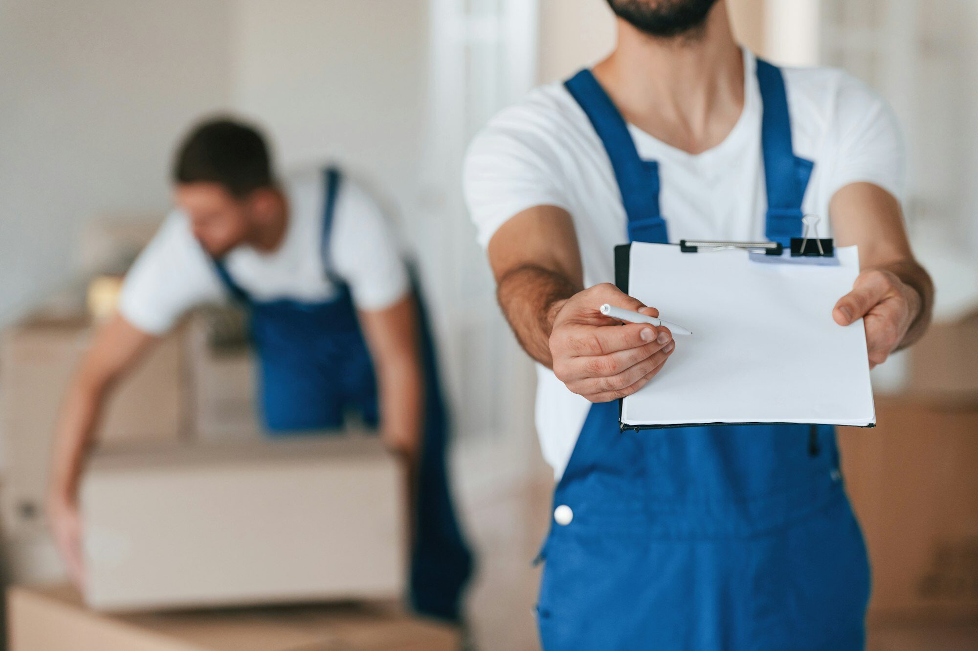 A man in blue overalls holds out a blank clipboard while another person organizes boxes in the background. The setting appears to be a moving or storage environment.