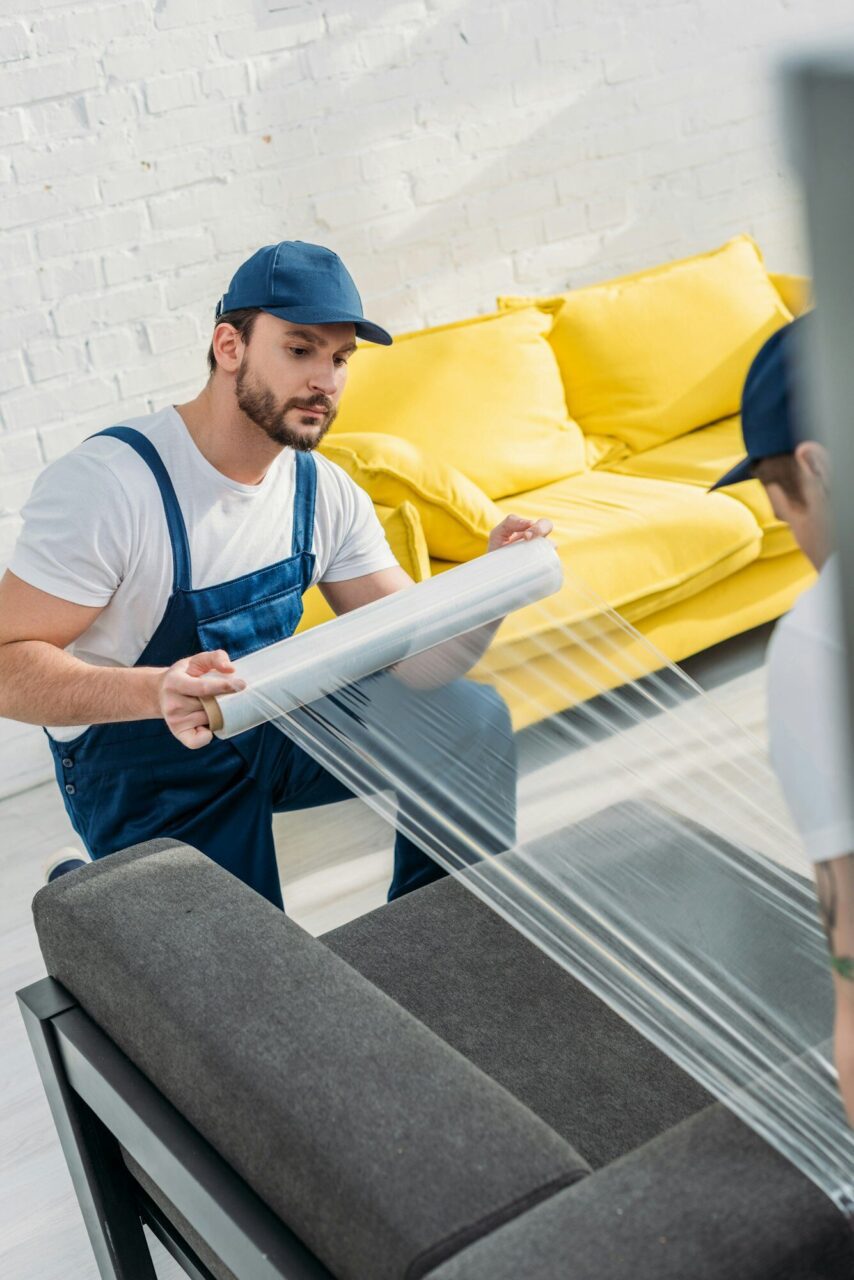 A worker in a blue cap and overalls is wrapping a dark sofa with plastic film, while a bright yellow couch is visible in the background. The setting appears to be a well-lit room with white walls.