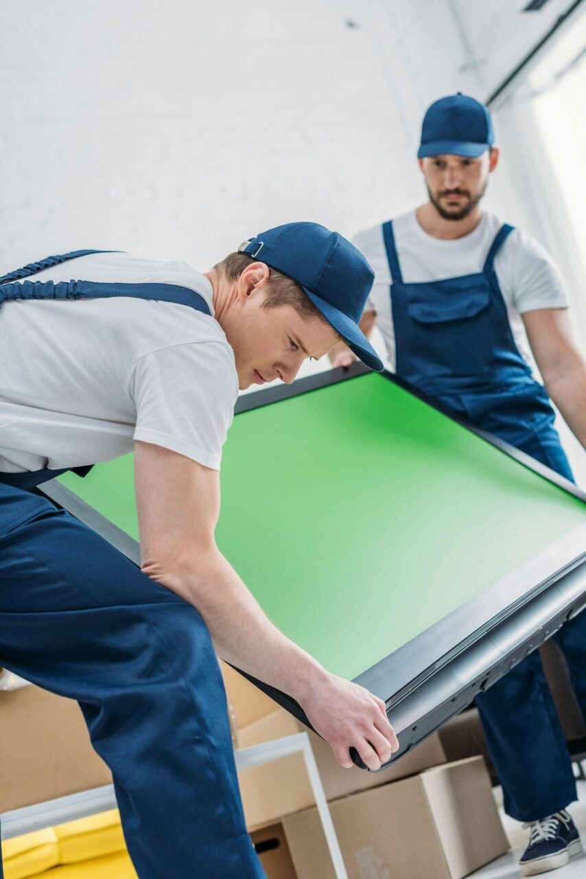 Two workers in blue overalls carefully lift a large green screen, preparing it for setup in a bright, spacious room. Boxes are visible in the background.