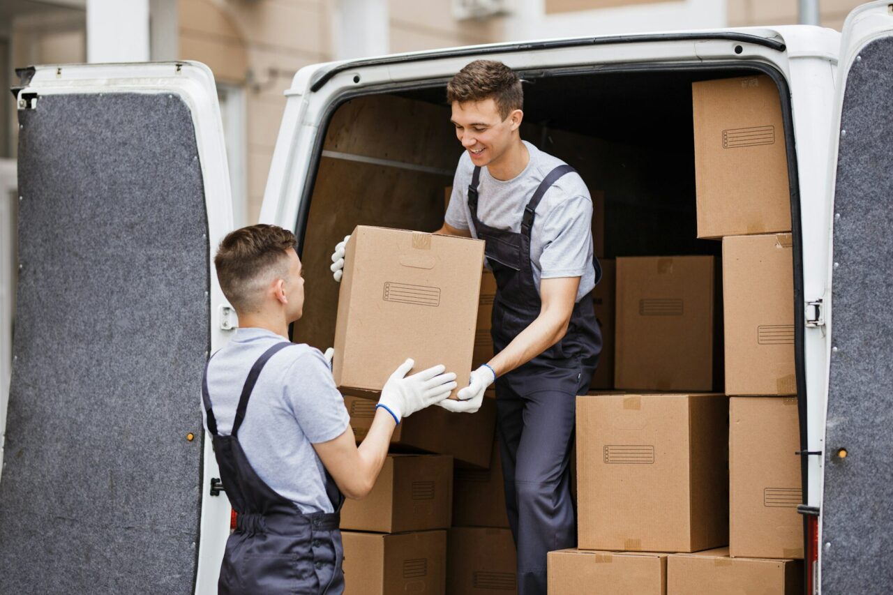 Two workers are loading cardboard boxes into a delivery van, collaborating efficiently. The scene depicts a busy logistics environment.