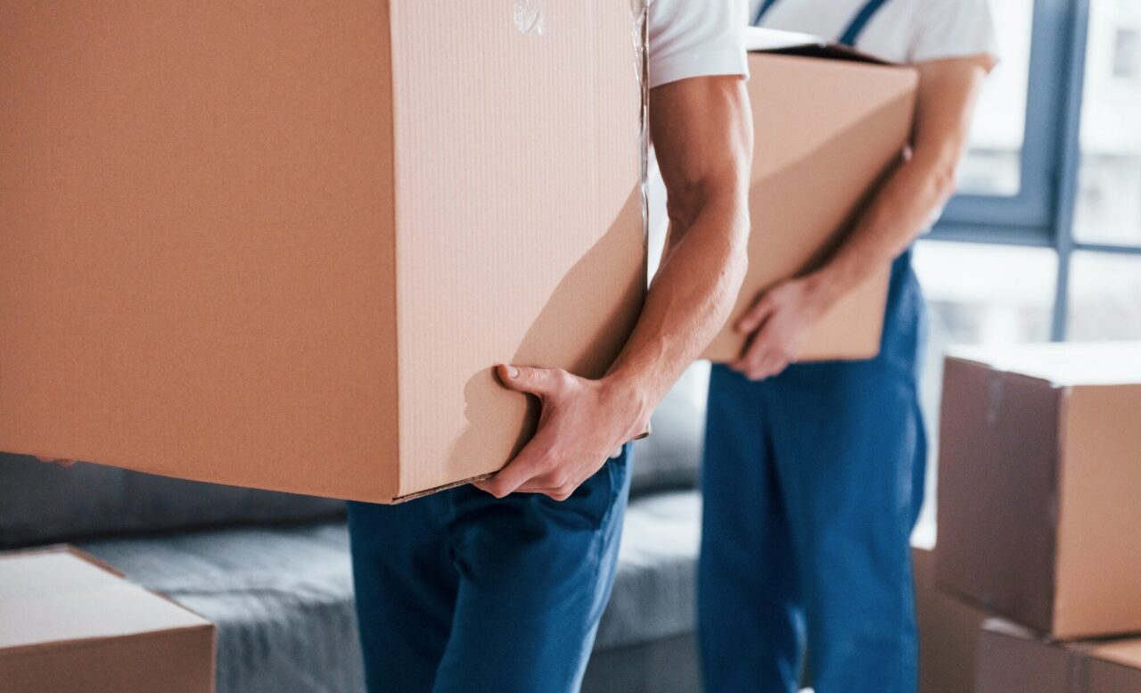Two individuals are carrying large cardboard boxes in a bright, modern room. The scene suggests a moving or unpacking process.