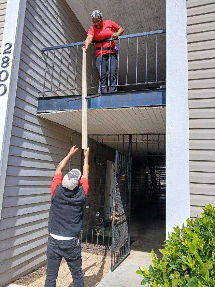 Two movers are coordinating the transfer of a long item from a balcony to the ground level, demonstrating teamwork and careful handling. The scene takes place in a residential area with a focus on efficient moving practices.