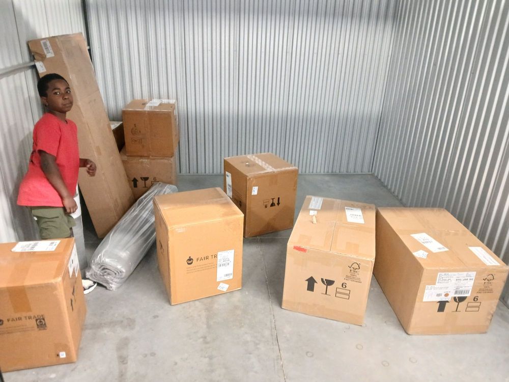 A young person stands beside several cardboard boxes in a storage unit, indicating preparation for a move. The setting is organized, showcasing the importance of careful packing and planning for relocation.